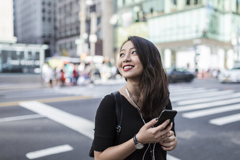USA, New York City, Manhattan, junge Frau hört Musik mit Handy und Kopfhörern auf der Straße - GIOF02489