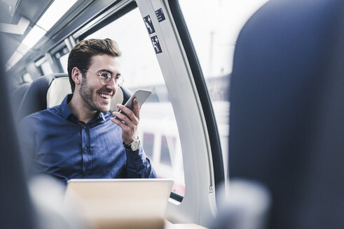 Glücklicher Geschäftsmann im Zug, der ein Mobiltelefon benutzt - UUF12633