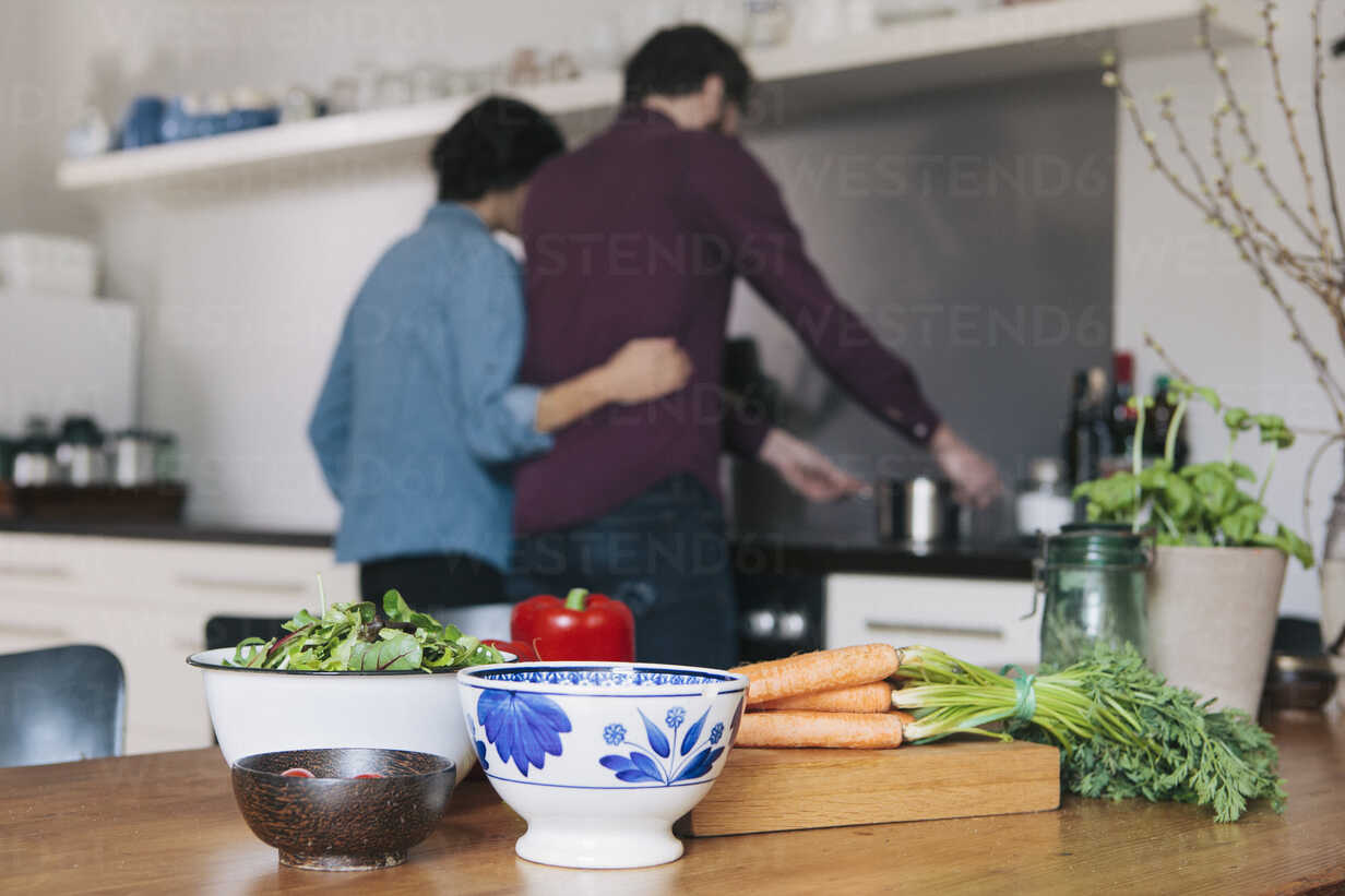 Vegetables On Kitchen Table With Couple Cooking In Background Stockphoto
