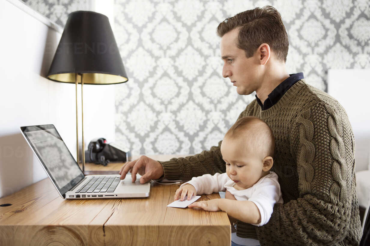 Father Working On Laptop Computer While Sitting With Son Table In Home Stockphoto