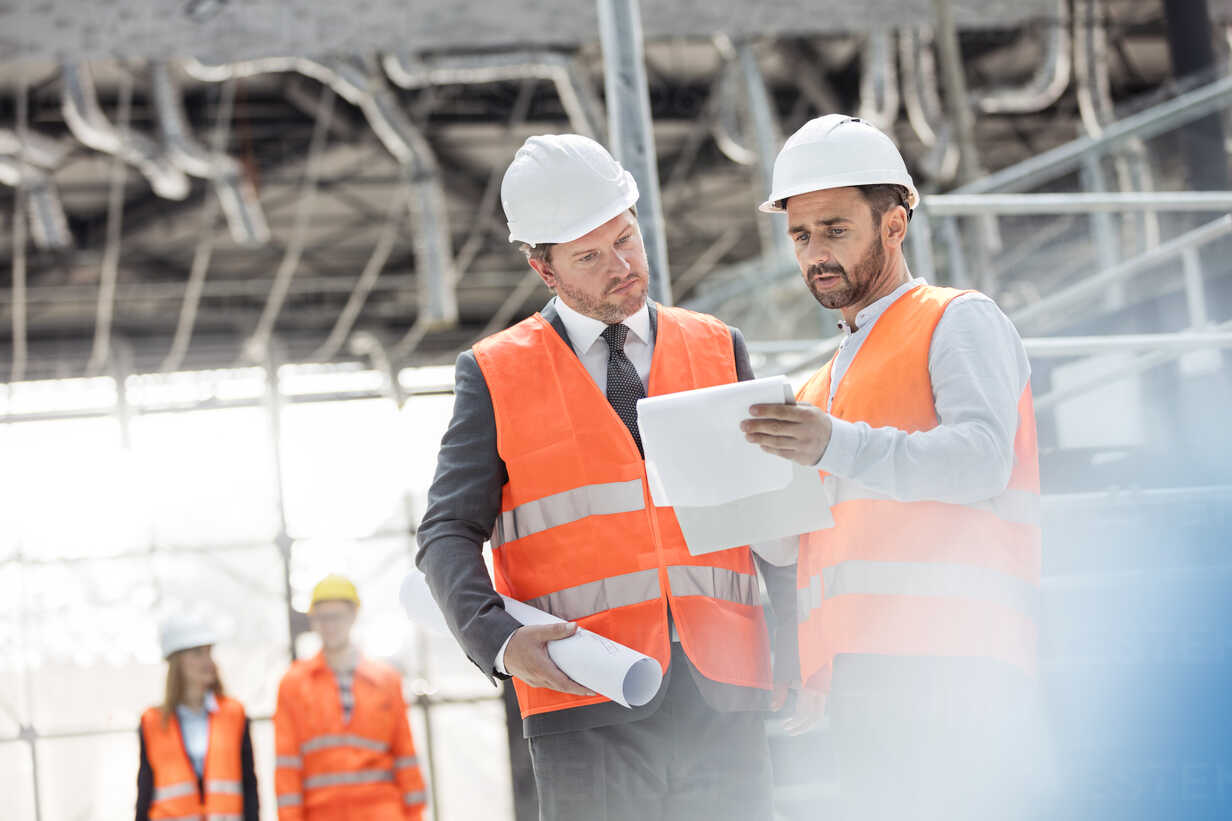 Male Engineers With Blueprints And Clipboard Discussing Paperwork At Construction Site Stockphoto