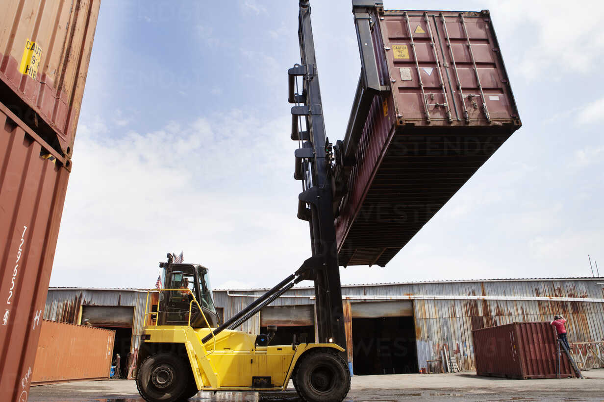 Man In Forklift Lifting Cargo Container At Commercial Dock Against Sky Stockphoto