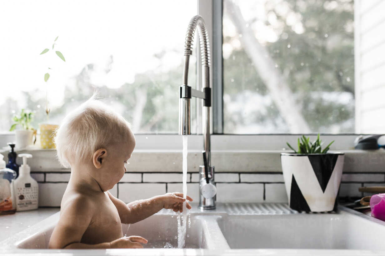 Side View Of Shirtless Toddler Playing With Water While Sitting In Kitchen Sink At Home Stockphoto
