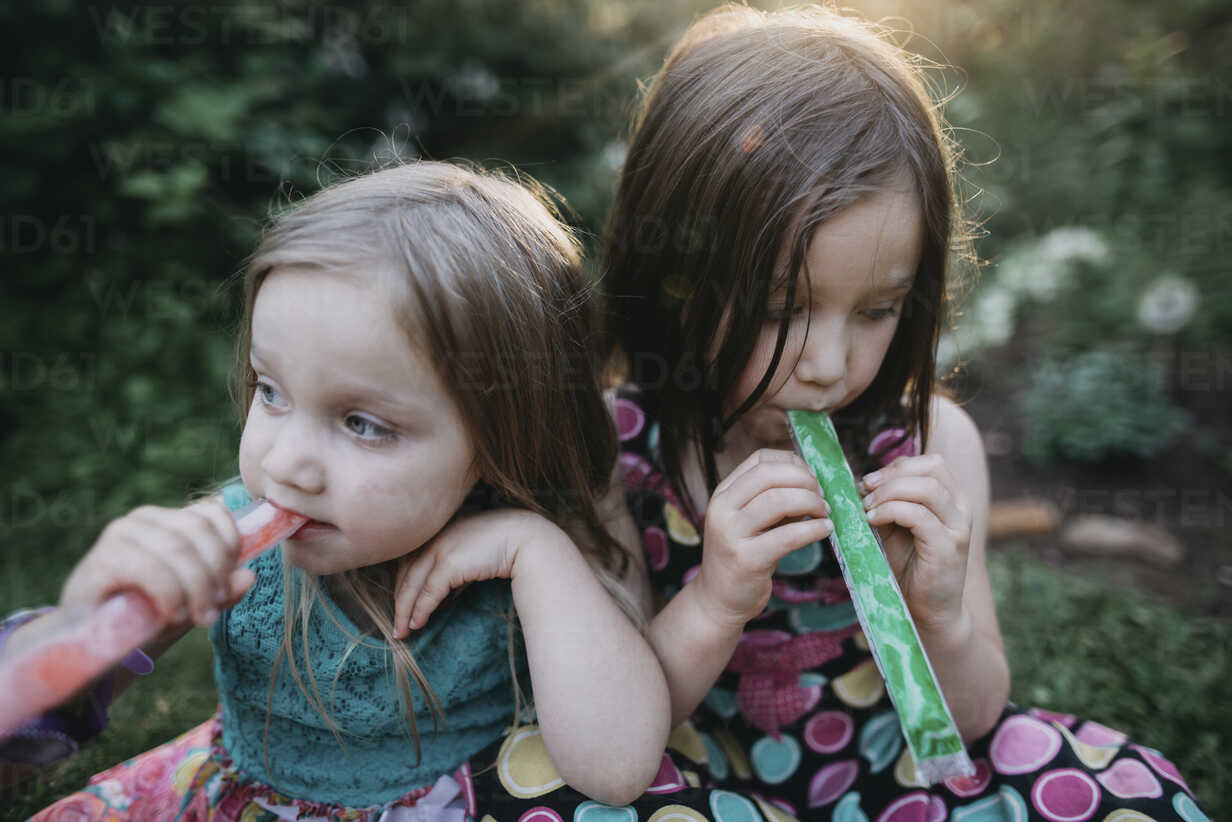 Sisters Looking Away While Sucking Plastic Tube Popsicles At Yard Stockphoto