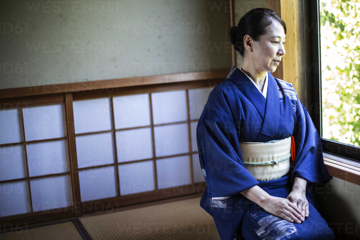 Japanese Woman Wearing Traditional Bright Blue Kimono With Cream Coloured Obi Kneeling On Floor In Traditional