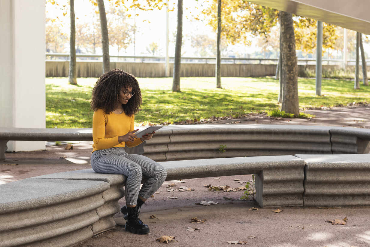 Afro Woman Using Digital Tablet While Sitting On Stone Bench In Public Park Stockphoto